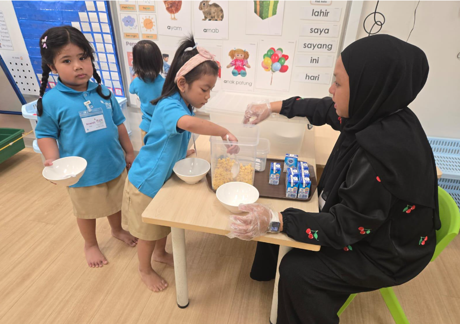 Teacher in hijab serves cereal and milk to barefoot children in blue uniforms at a classroom table.